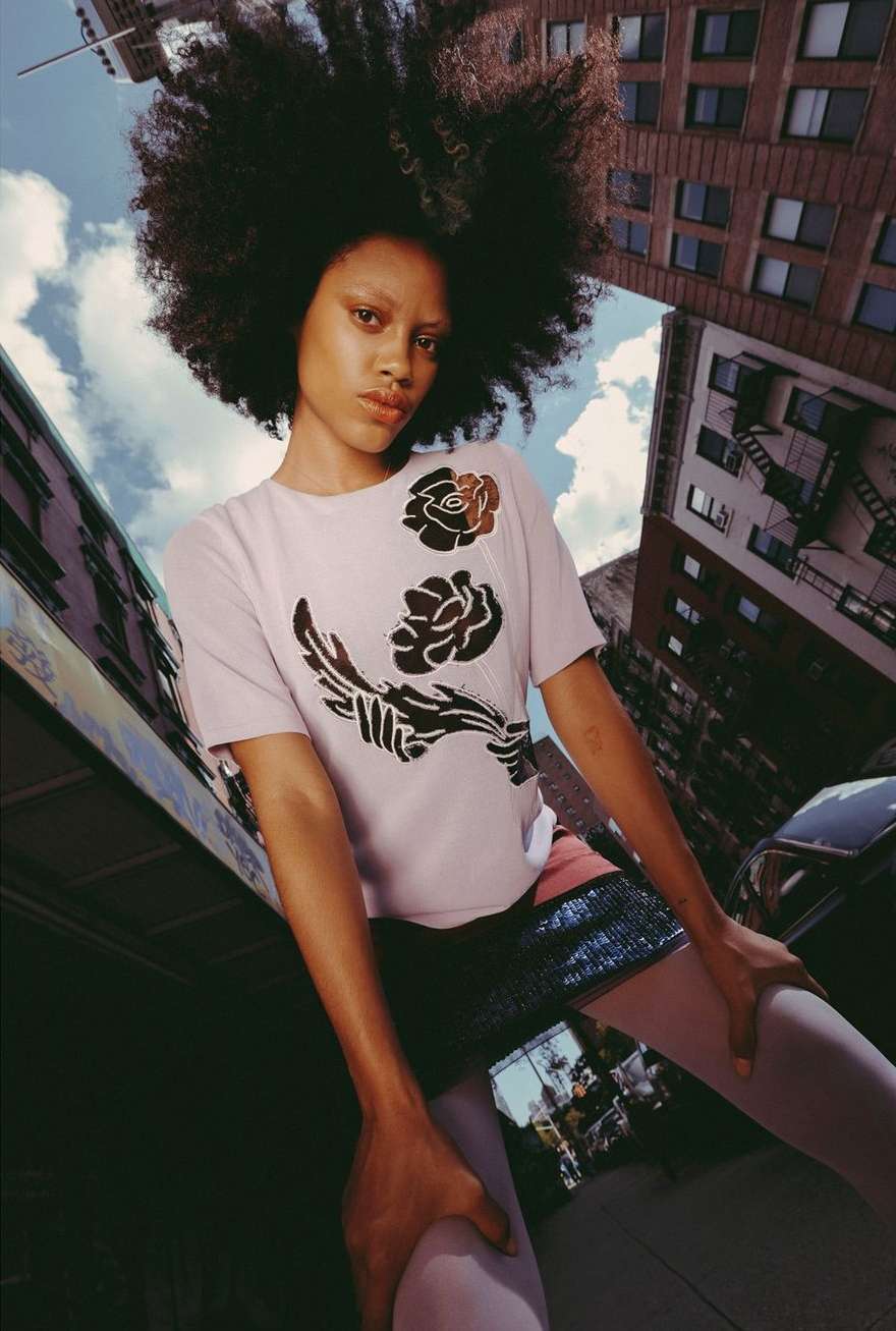 Eye-level view of woman with afro wearing a floral t-shirt in urban setting surrounded by city buildings and partly cloudy sky.