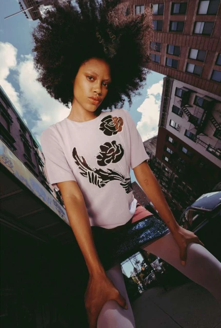 Eye-level view of woman with afro wearing a floral t-shirt in urban setting surrounded by city buildings and partly cloudy sky.