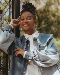 Smiling Black woman with locs and hoop earrings wearing a denim jacket and white shirt outdoors by a fence, photographed by Adrienne Raquel.