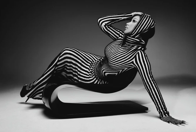 Woman in a black and white striped hooded jumpsuit reclining on a curved modern chair against a grey studio backdrop.