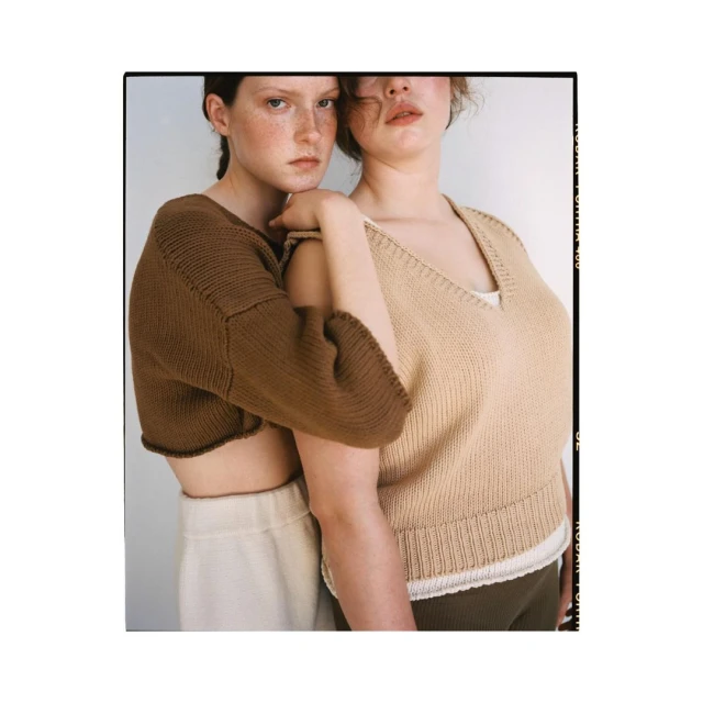 Two fair-skinned young adult women pose indoors wearing knit tops and neutral-toned pants against a light neutral background in a studio shot.