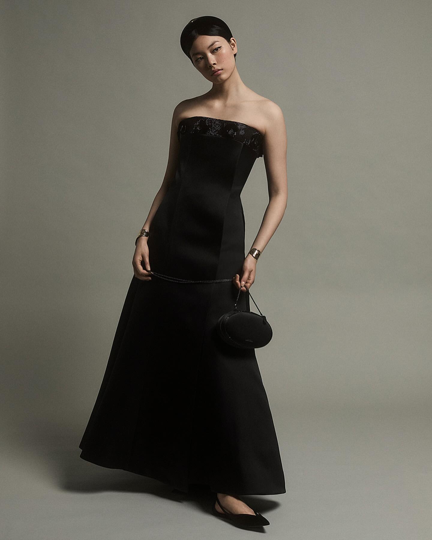 Woman in a black strapless evening dress holding a small handbag against a solid grey background