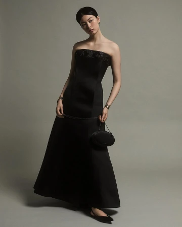 Woman in a black strapless evening dress holding a small handbag against a solid grey background