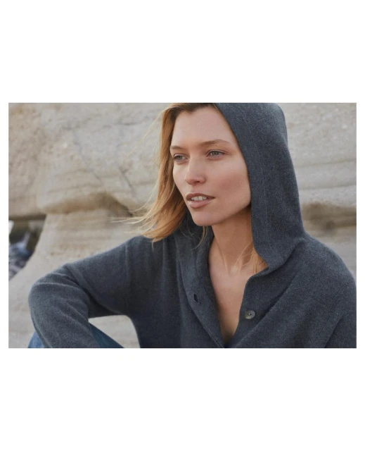 Blonde woman in a hooded sweater sitting outdoors with a neutral facial expression and light brown hair in a medium shot.