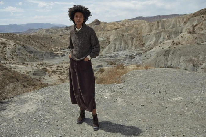 Model with dark curly hair wearing a brown knit sweater and long skirt standing in a rugged mountainous desert landscape.