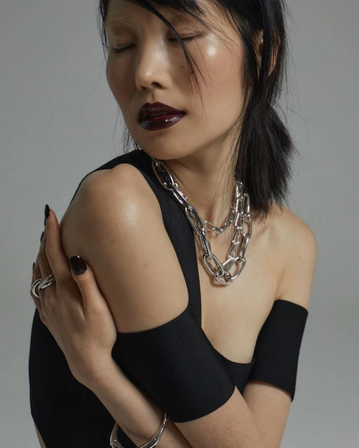 Asian woman with glossy dark lipstick and silver necklace in a studio. She is wearing a black top and silver ring and bangle.