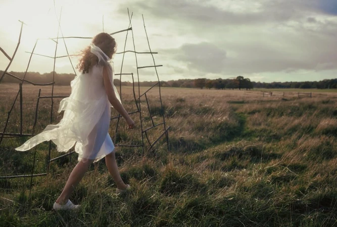 Woman in white dress and sheer veil walking through a grassy field past a rustic stick structure at sunset by Agata Pospieszynska.