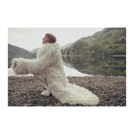 Adult female poses lakeside wearing long white faux fur coat. Background features calm lake and forested mountains on overcast day.