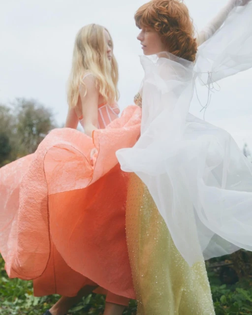 Two fair-skinned women in flowing dresses stand outside; one in peach, the other in layered white fabric in a fashion editorial.