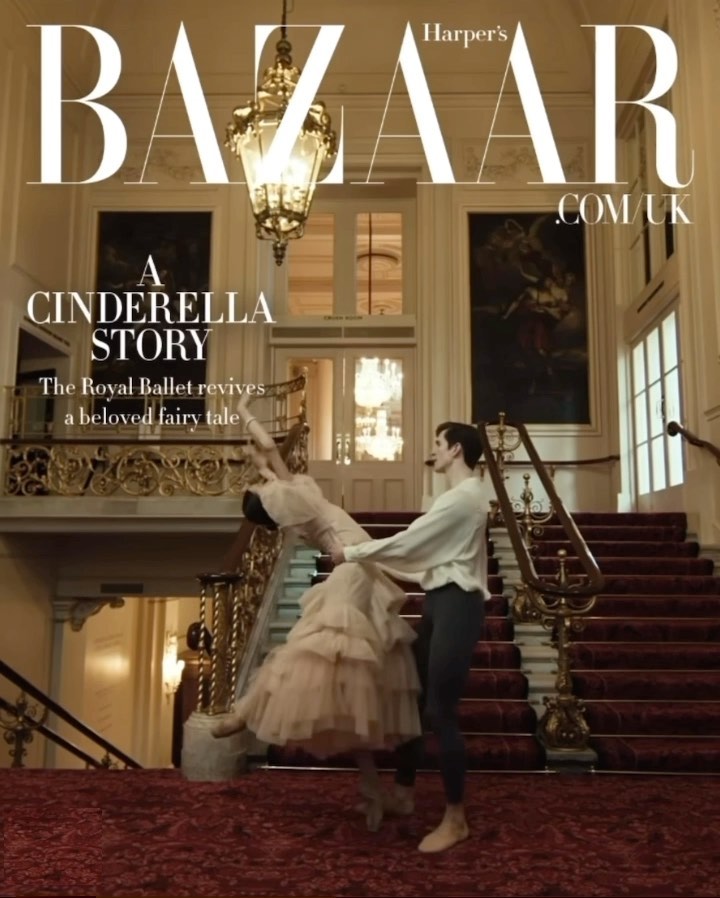 A couple of ballet dancers are posed on a staircase indoors in front of a large chandelier in an editorial fashion photograph.