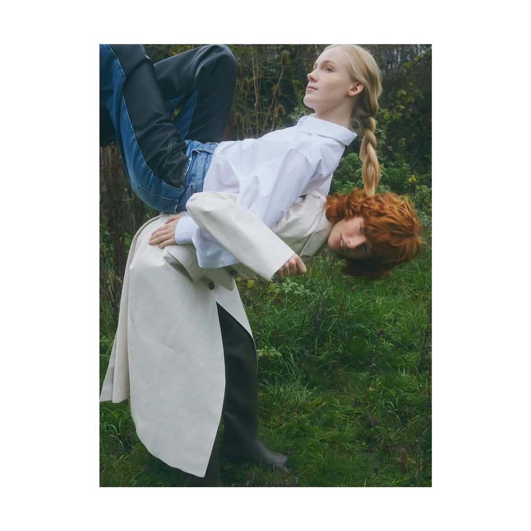 Two women in a forest setting. One woman, in jeans and a white shirt, is held up by the other. The scene has a fashion editorial aesthetic.