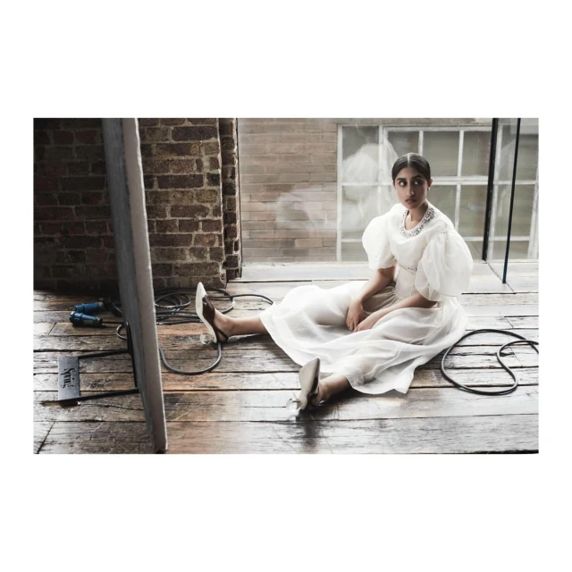 Woman in a sheer white dress poses on a hardwood floor near a brick wall with window light, accented by studio equipment and natural light.