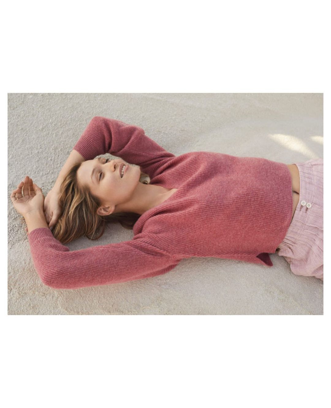 Woman in a rose-colored sweater and light pink pants is lying on her back on a textured light beige surface, with her hands near her head.