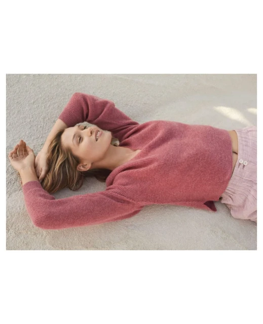 Woman in a rose-colored sweater and light pink pants is lying on her back on a textured light beige surface, with her hands near her head.