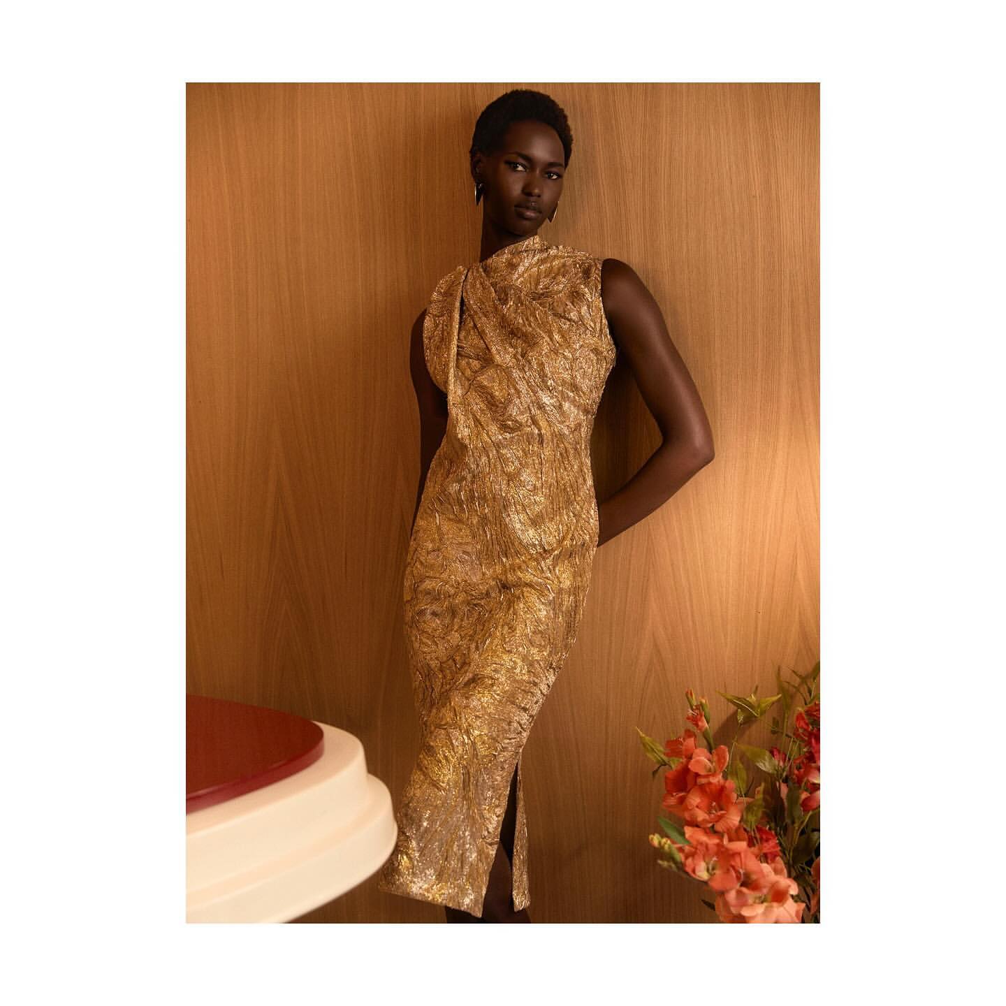 Elegant adult woman in a sleeveless golden dress posing against a warm wooden backdrop, accented by flowers and a side table.
