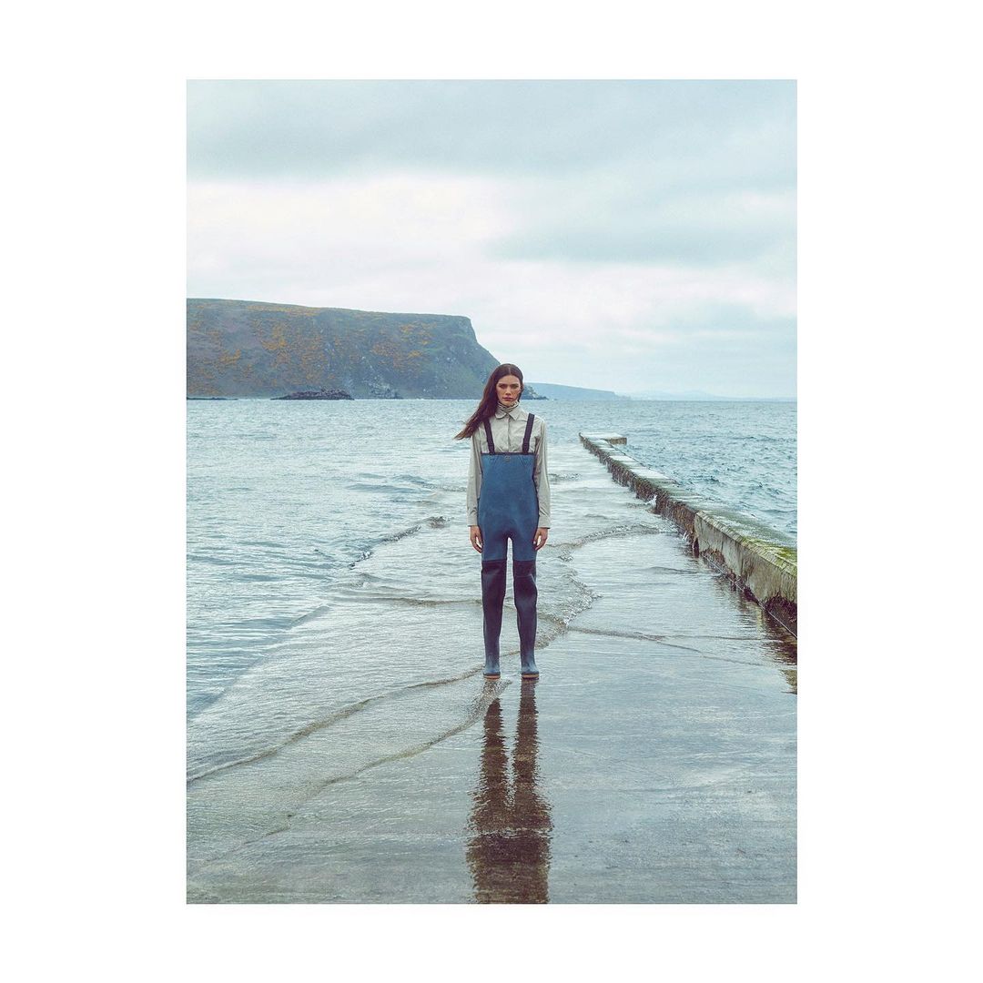 Woman stands on a pier wearing blue overalls and boots in a landscape with a cloudy sky, sea, and distant land.