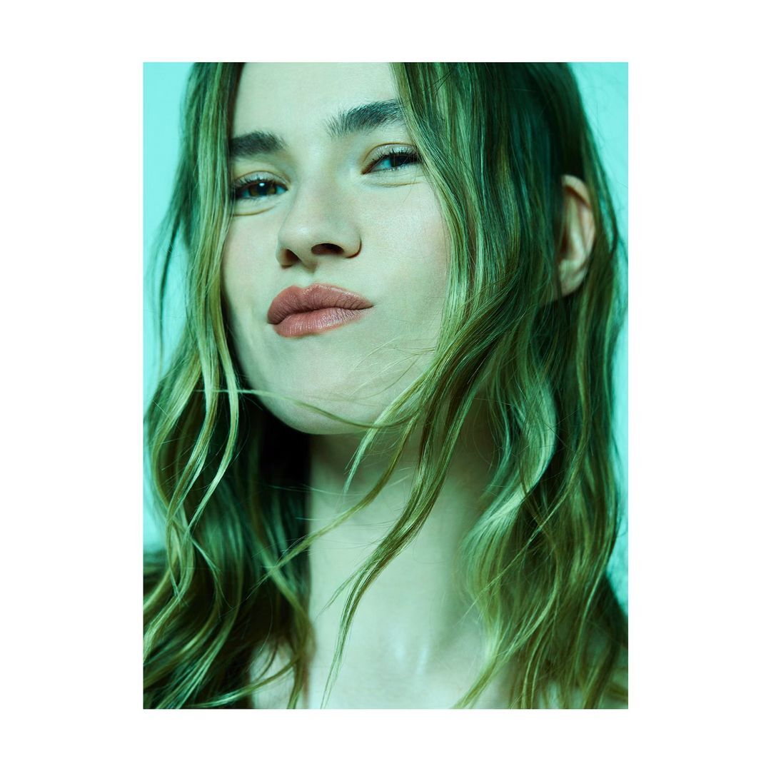 Close-up portrait of a young adult female with long, blonde wavy hair and fair skin in front of a light green background.