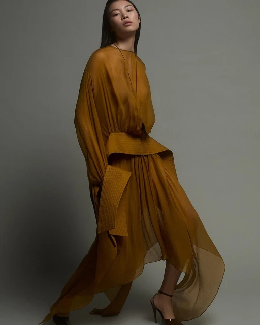 A fashionable Asian woman poses in a flowing yellow dress and heels against a neutral background in a studio fashion shot.
