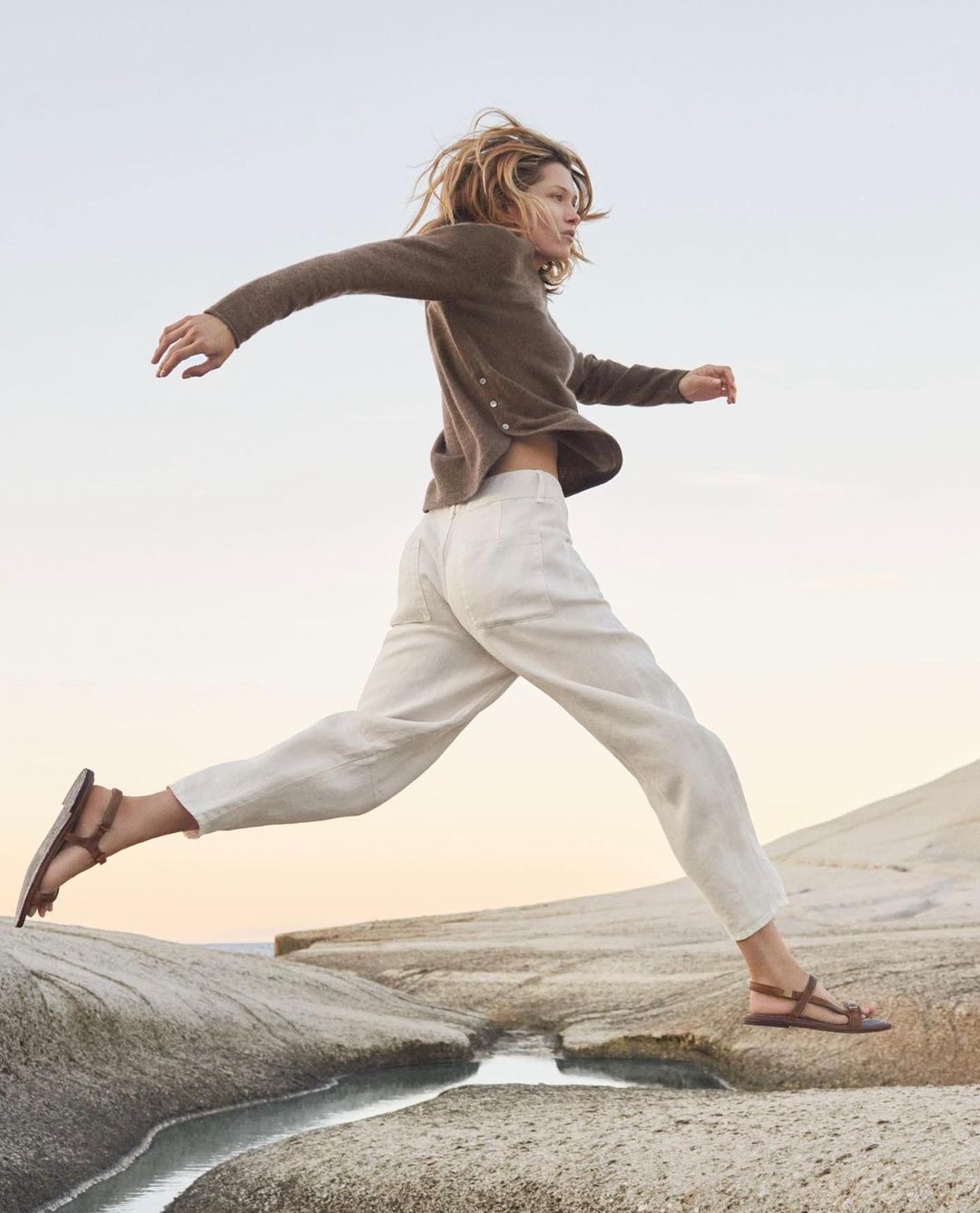 An adult female leaps between rock formations on a beach at dusk, wearing casual clothing: a cardigan, pants, and sandals, with a blurred sky.