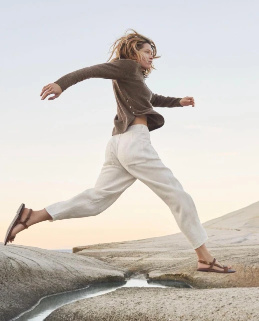 An adult female leaps between rock formations on a beach at dusk, wearing casual clothing: a cardigan, pants, and sandals, with a blurred sky.