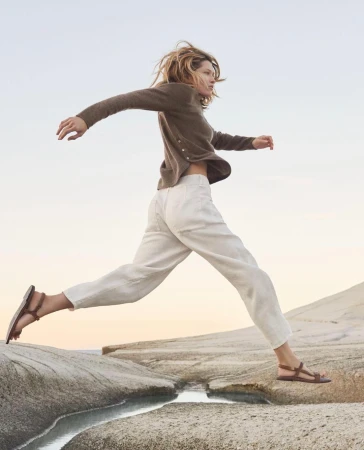 An adult female leaps between rock formations on a beach at dusk, wearing casual clothing: a cardigan, pants, and sandals, with a blurred sky.