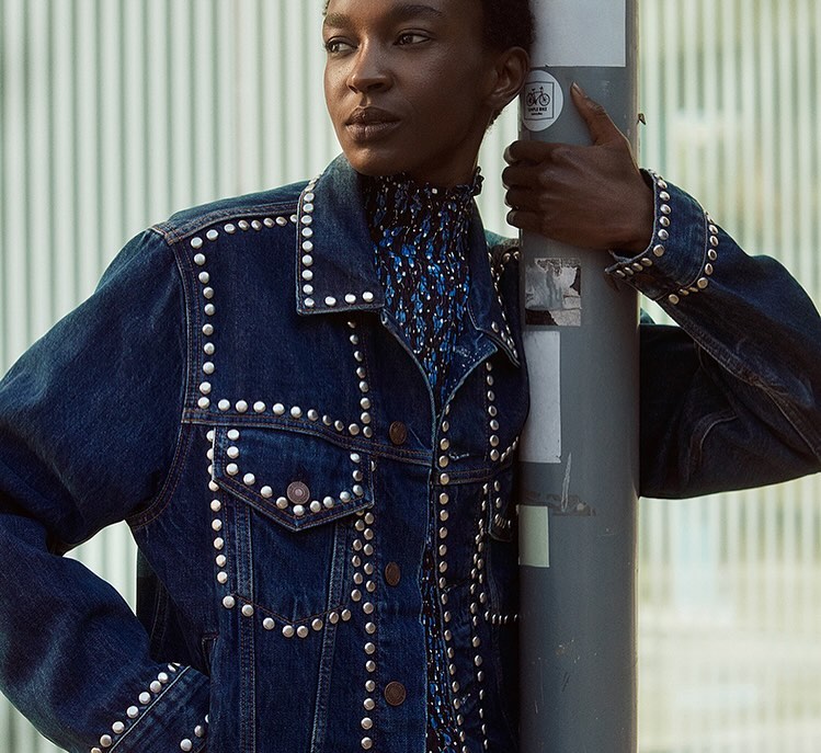 Adult female model in denim jacket with silver studs and a blue patterned top, posing outdoors in an urban setting, medium shot.