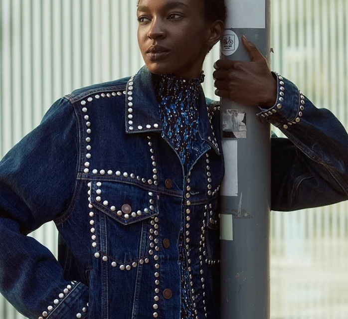 Adult female model in denim jacket with silver studs and a blue patterned top, posing outdoors in an urban setting, medium shot.