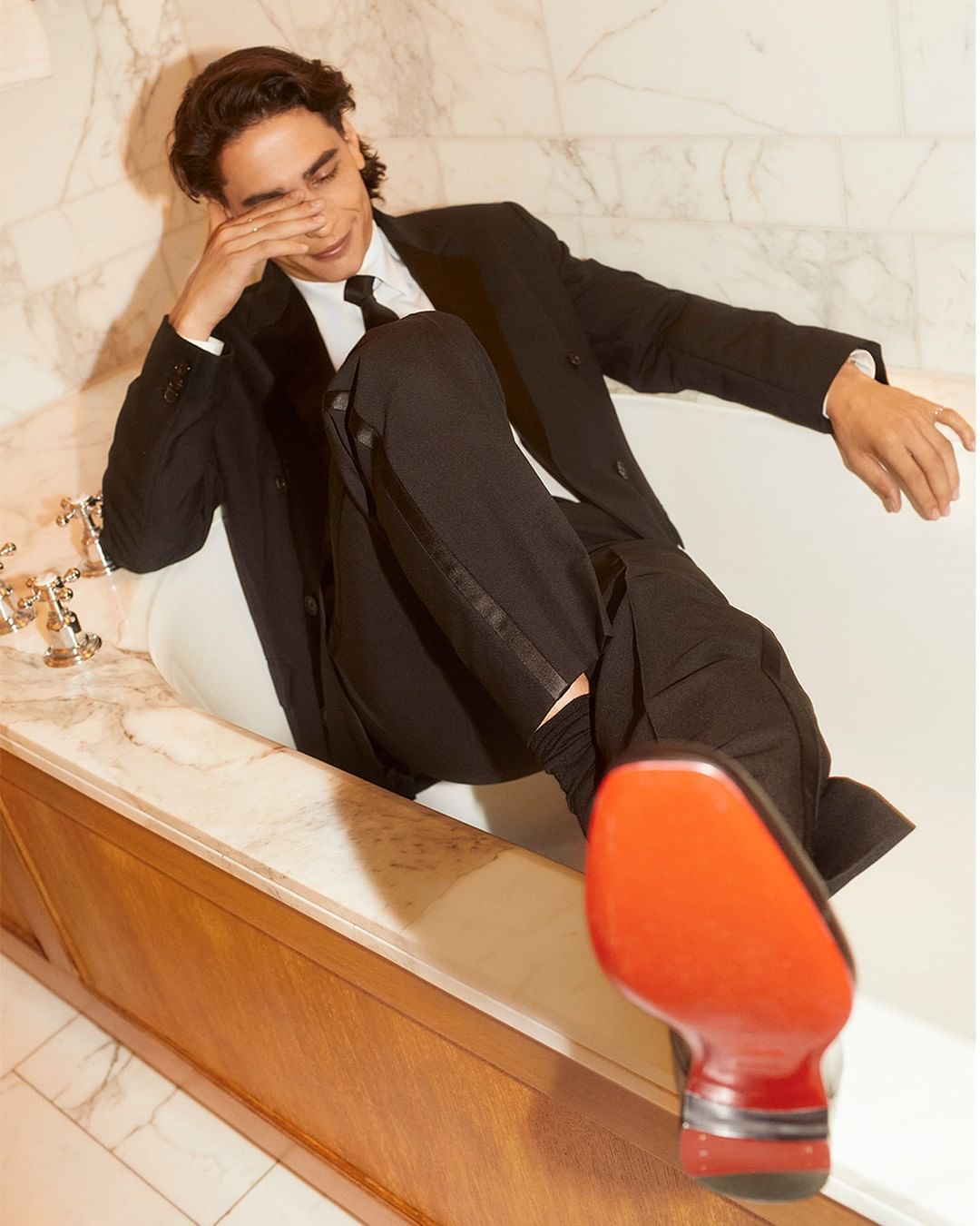 Adult male fashion model in black suit, posed in a bright white bathtub with red bottom dress shoe. Relaxed pose, indoor setting.