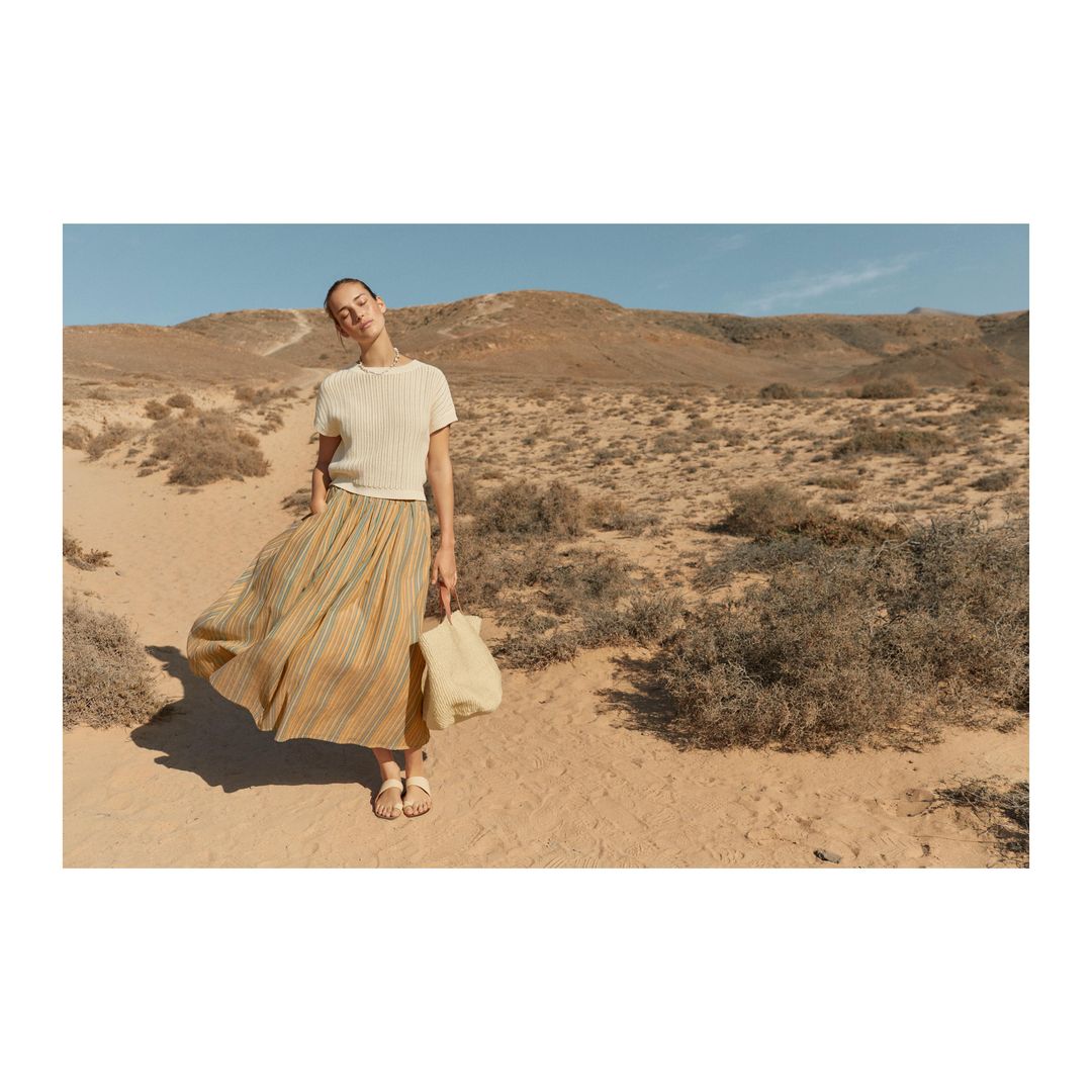 A woman in a light-colored sweater and striped skirt stands in a desert landscape holding a tote bag, bathed in warm sunlight.
