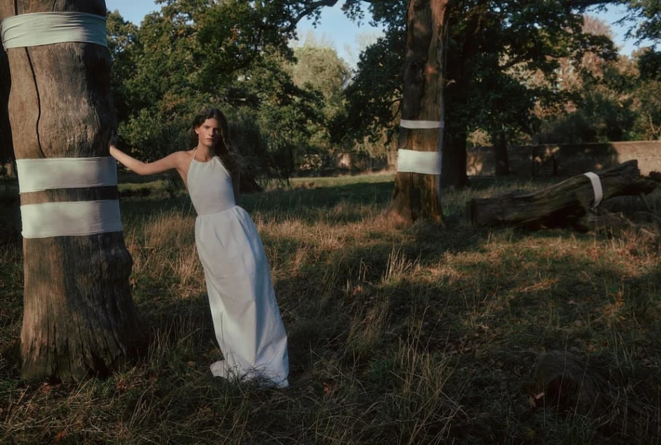 A woman in a white dress leans against a tree with a white sash in a grassy forest clearing on a sunny day.