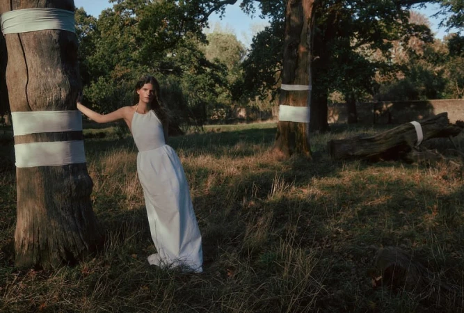 A woman in a white dress leans against a tree with a white sash in a grassy forest clearing on a sunny day.