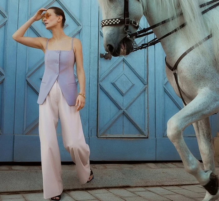 Stylish woman in lilac top and wide-leg trousers stands with white horse by blue door, fashion photography, outdoor setting.