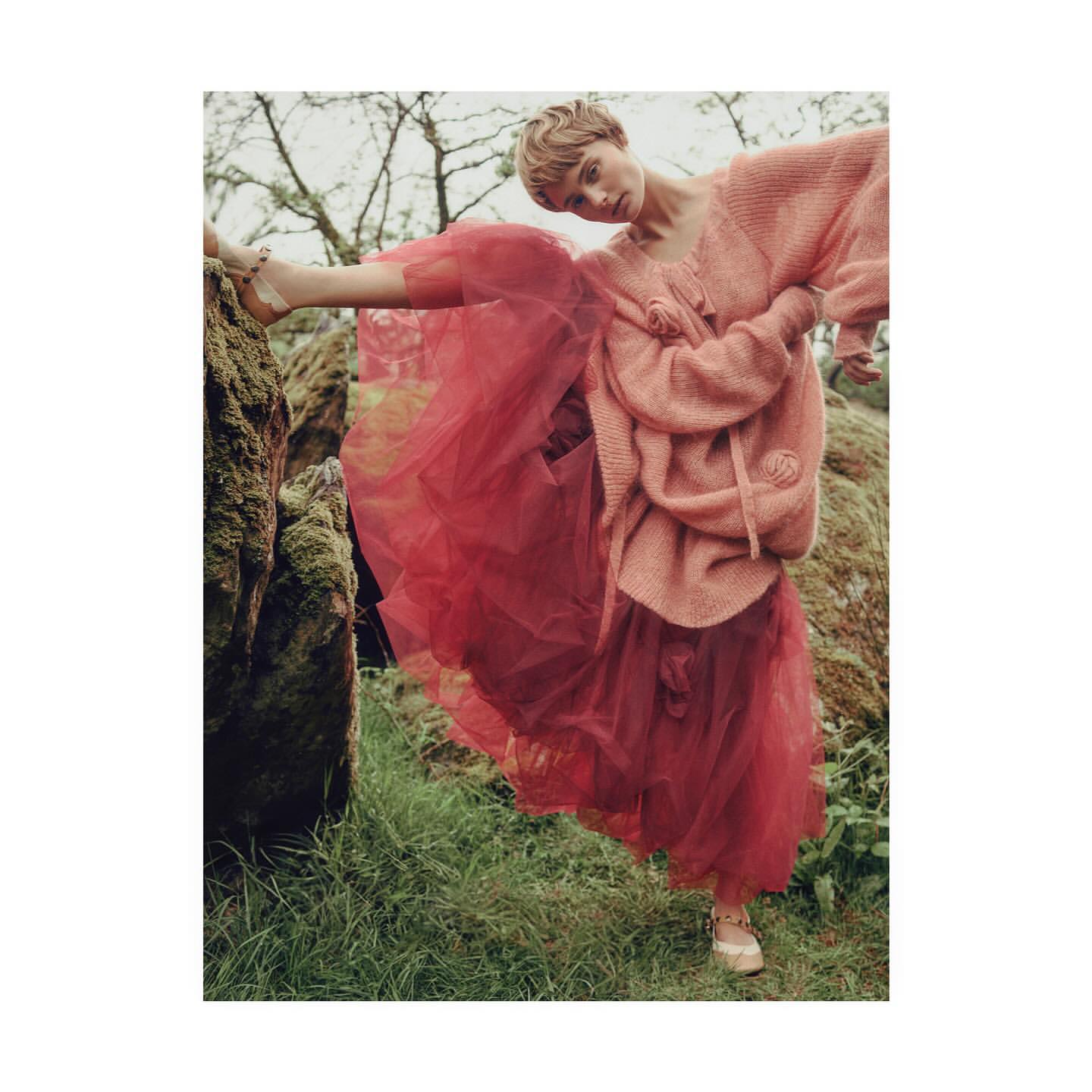 A fashionable young woman with short blonde hair poses outdoors in a pink sweater and red tulle skirt near a moss-covered rock.