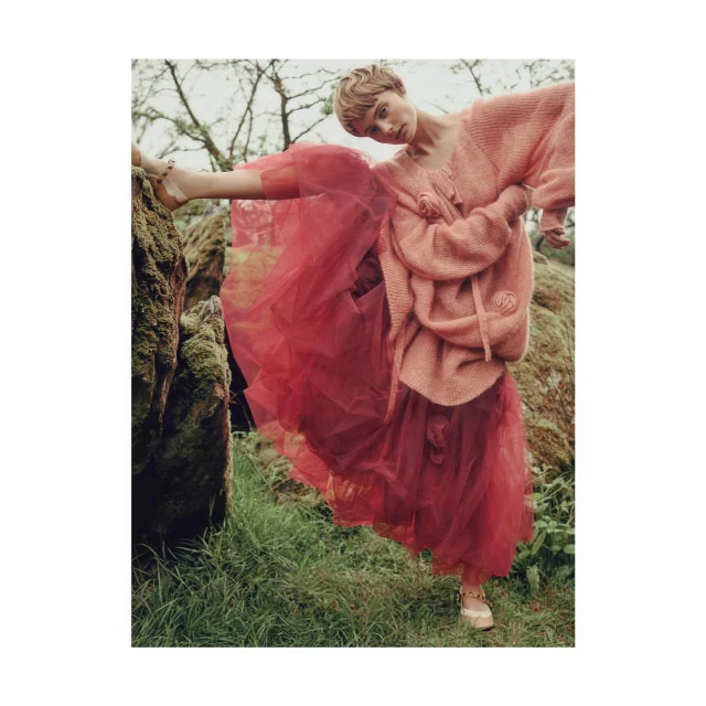 A fashionable young woman with short blonde hair poses outdoors in a pink sweater and red tulle skirt near a moss-covered rock.