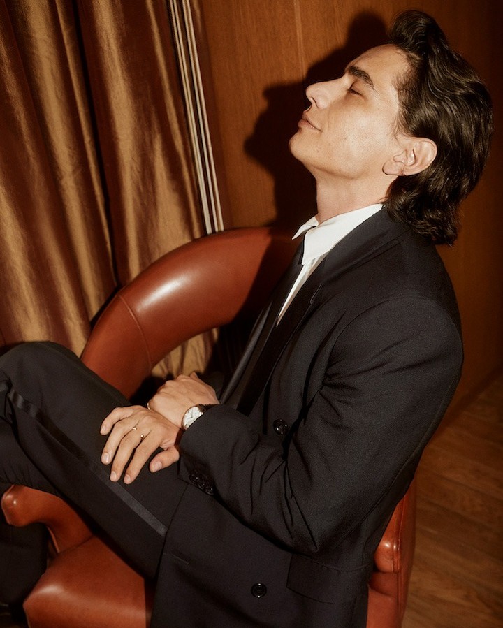 Portrait of a man wearing a black suit, seated in a leather chair against a warm-toned wall, with his eyes closed in a relaxed pose.