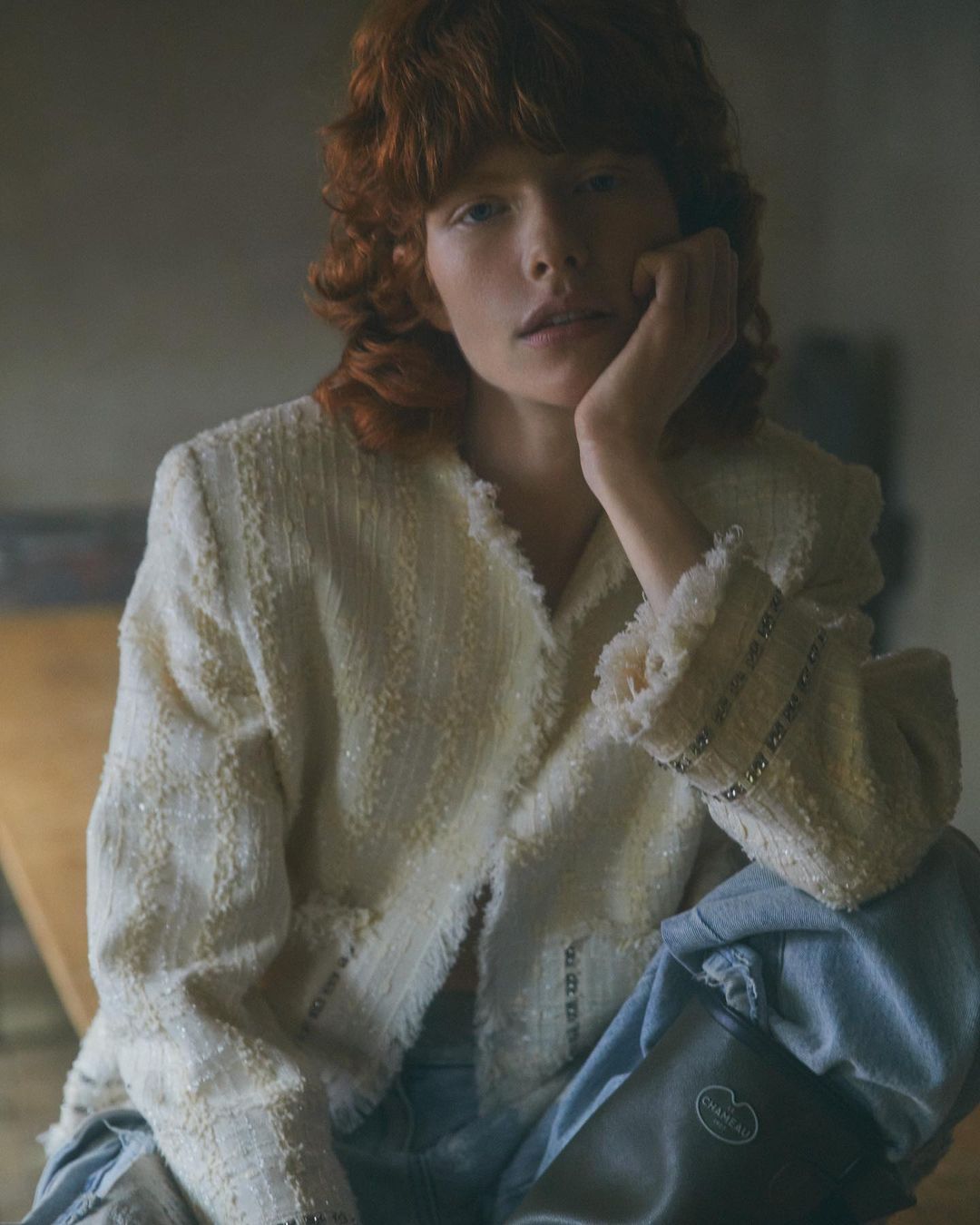 Redhead woman wearing a cream-colored boucle jacket and light blue jeans sits in a dimly lit room, resting her head on her hand.
