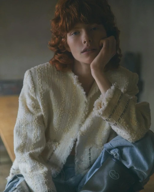Redhead woman wearing a cream-colored boucle jacket and light blue jeans sits in a dimly lit room, resting her head on her hand.
