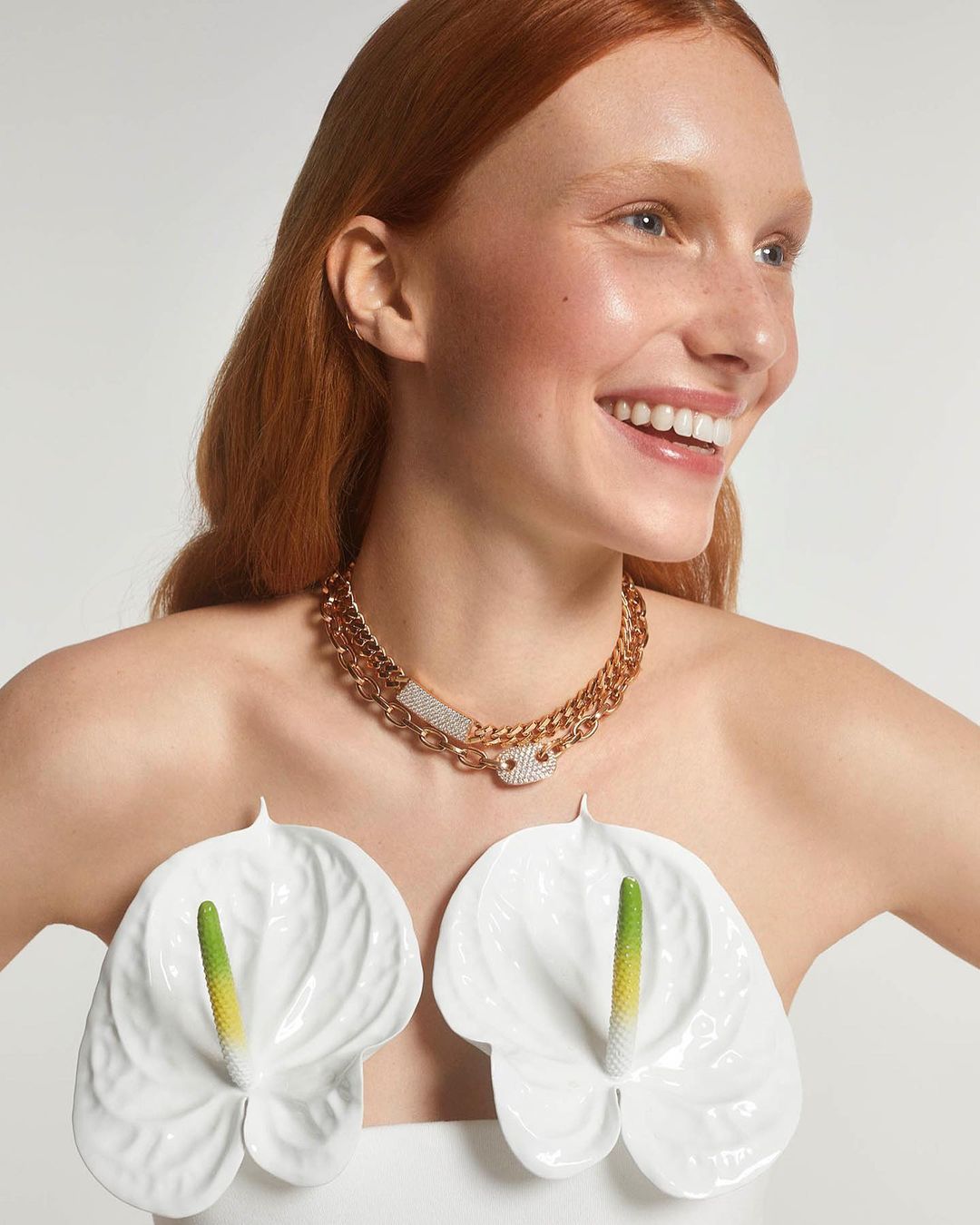 Portrait of a smiling, fair-skinned woman with red hair wearing a gold chain necklace and white lily flowers against a white background.