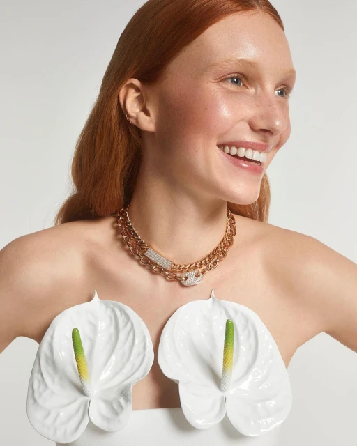 Portrait of a smiling, fair-skinned woman with red hair wearing a gold chain necklace and white lily flowers against a white background.