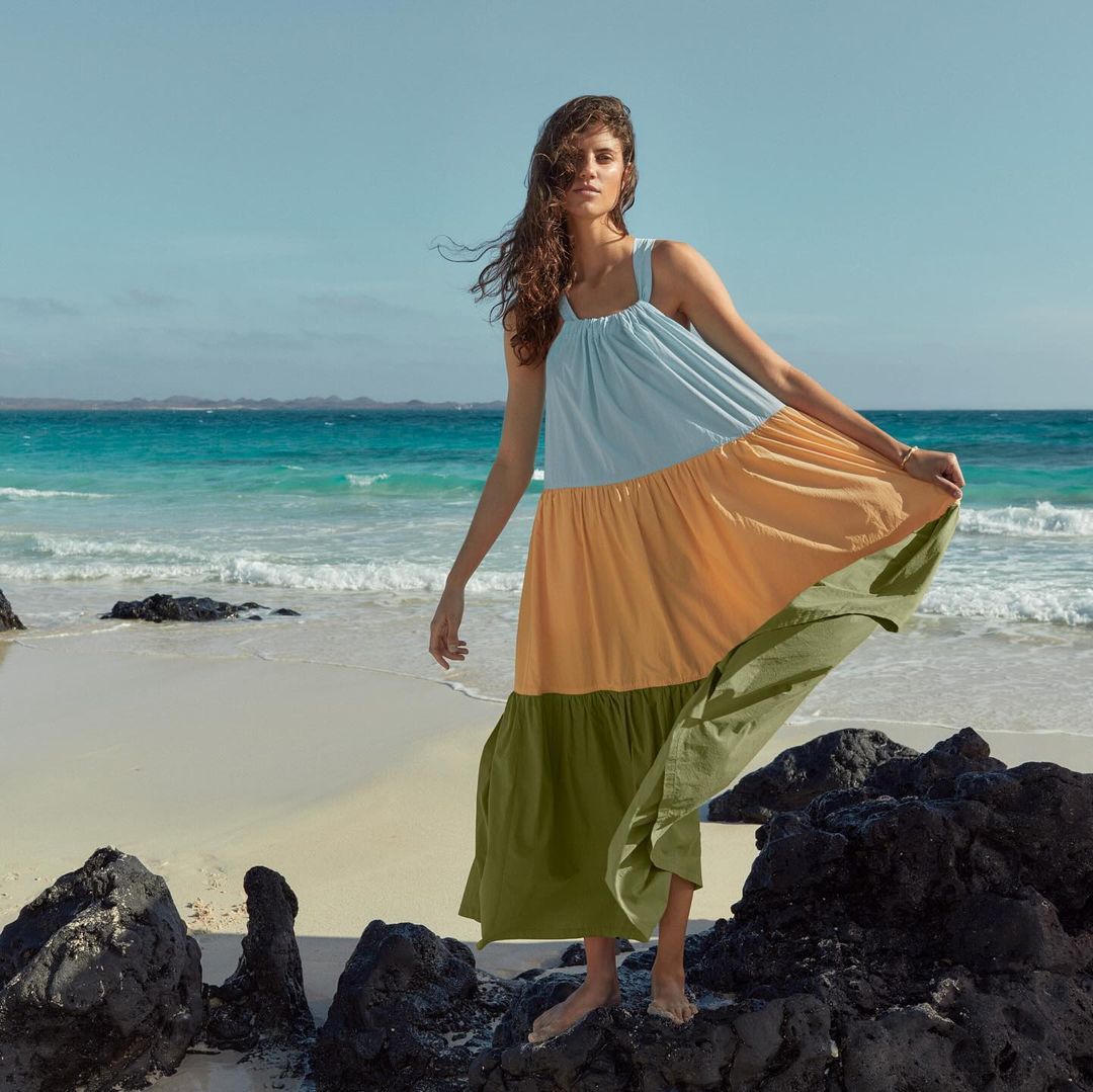 A woman in a flowy summer dress stands on a rocky beach with ocean and blue sky in the background, enjoying the sunny weather.