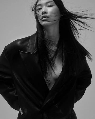 Portrait of an East Asian woman with long dark hair blowing in the wind, wearing a black leather blazer against a plain light background