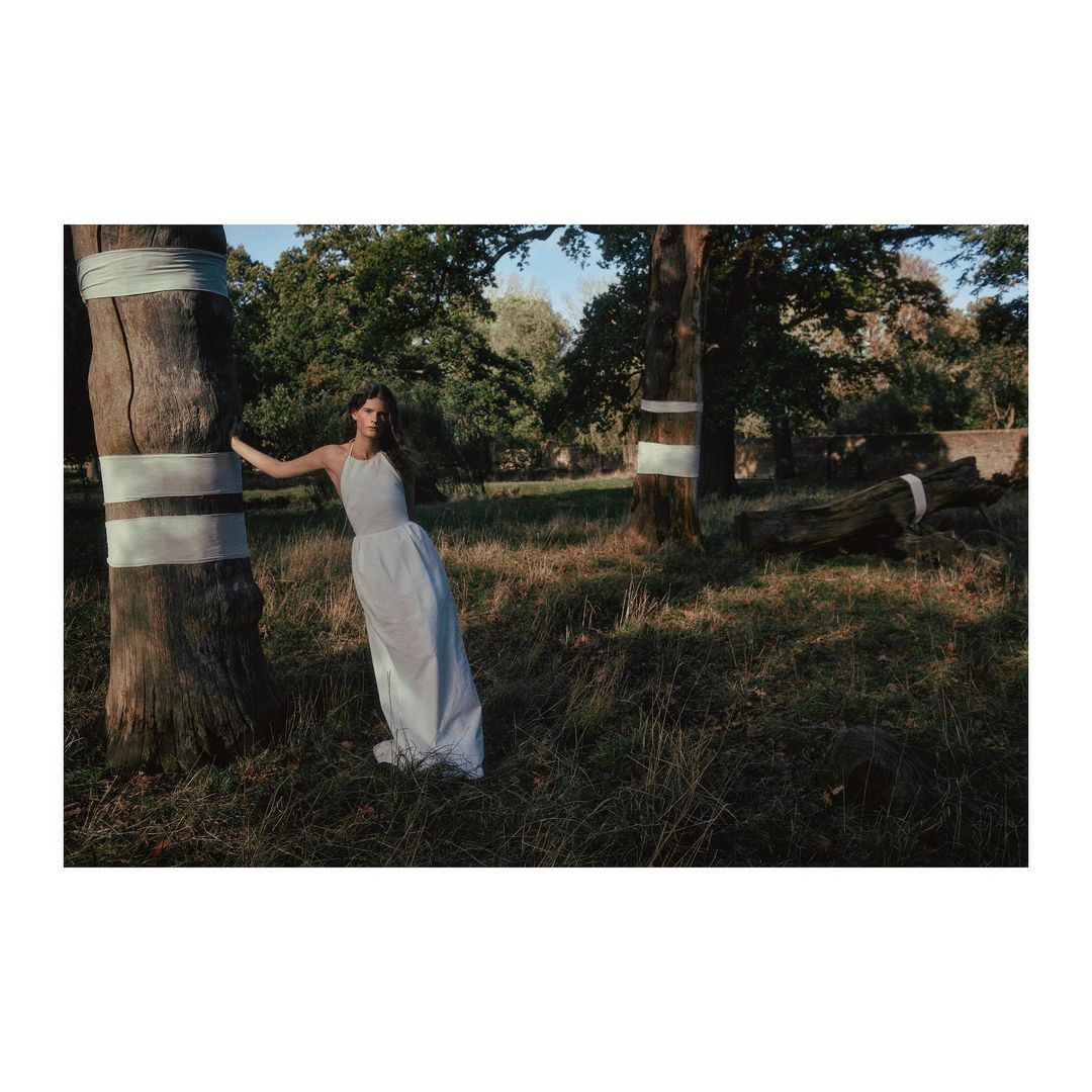 A woman in a white dress leans against a tree in a sunlit forest clearing, creating a serene and ethereal atmosphere in a natural setting.