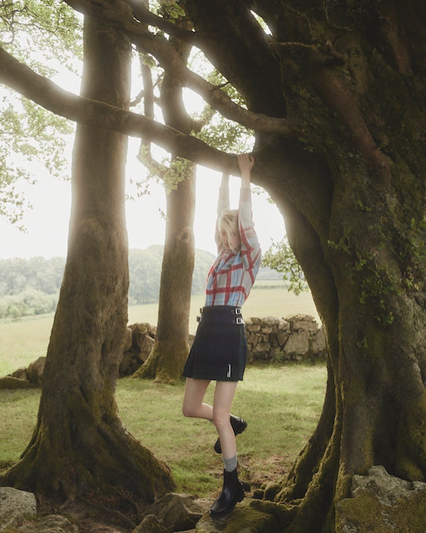 Young woman in a plaid top and dark skirt hangs from a tree branch in a lush, green field; a blend of fashion and nature.