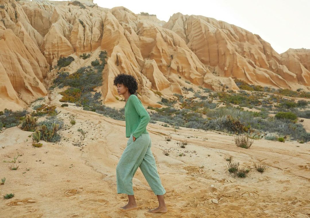 A young woman in casual, light-green clothing walks barefoot in a desert landscape with towering sandstone formations in the background.