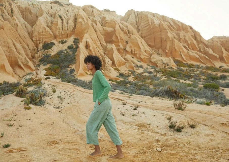 A young woman in casual, light-green clothing walks barefoot in a desert landscape with towering sandstone formations in the background.