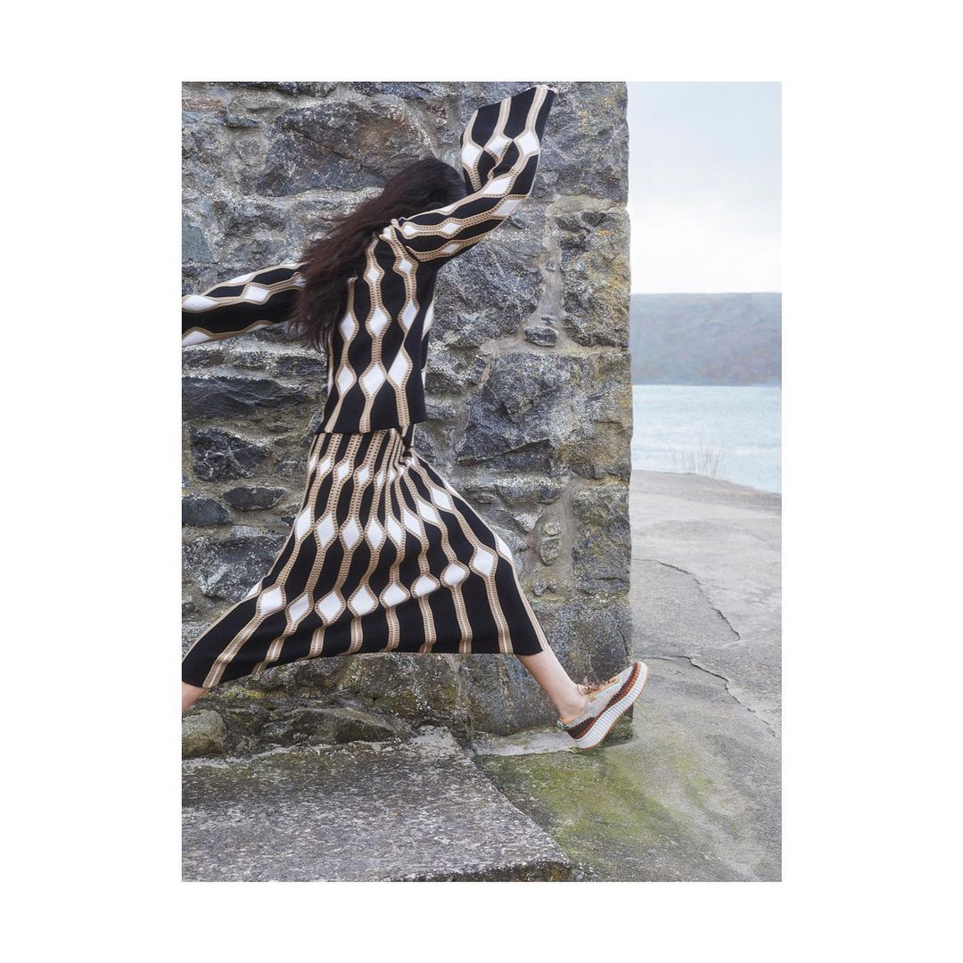 Woman in patterned dress leaps near stone wall with a water view backdrop. Fashion editorial photo, natural light, elegant outdoor setting.