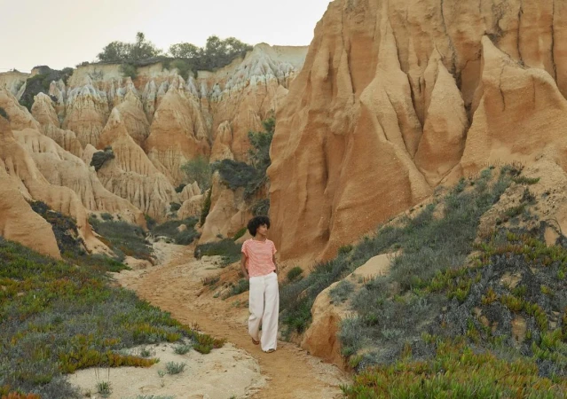 A woman walks along a sandy path through a canyon with eroded cliffs, wearing white pants and a peach-colored shirt in natural light.