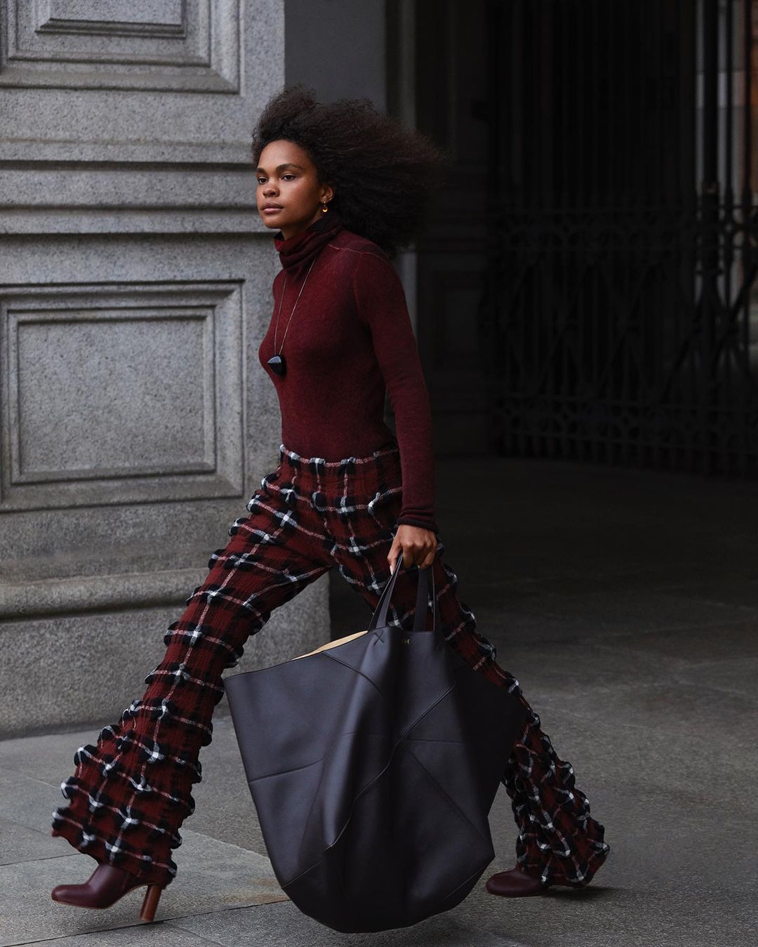 A woman walks outdoors holding a large tote bag. She wears flared, plaid pants, a turtleneck top, and heeled booties in a street setting.