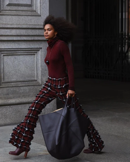 A woman walks outdoors holding a large tote bag. She wears flared, plaid pants, a turtleneck top, and heeled booties in a street setting.
