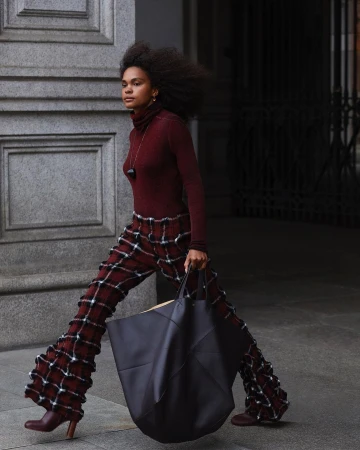 A woman walks outdoors holding a large tote bag. She wears flared, plaid pants, a turtleneck top, and heeled booties in a street setting.